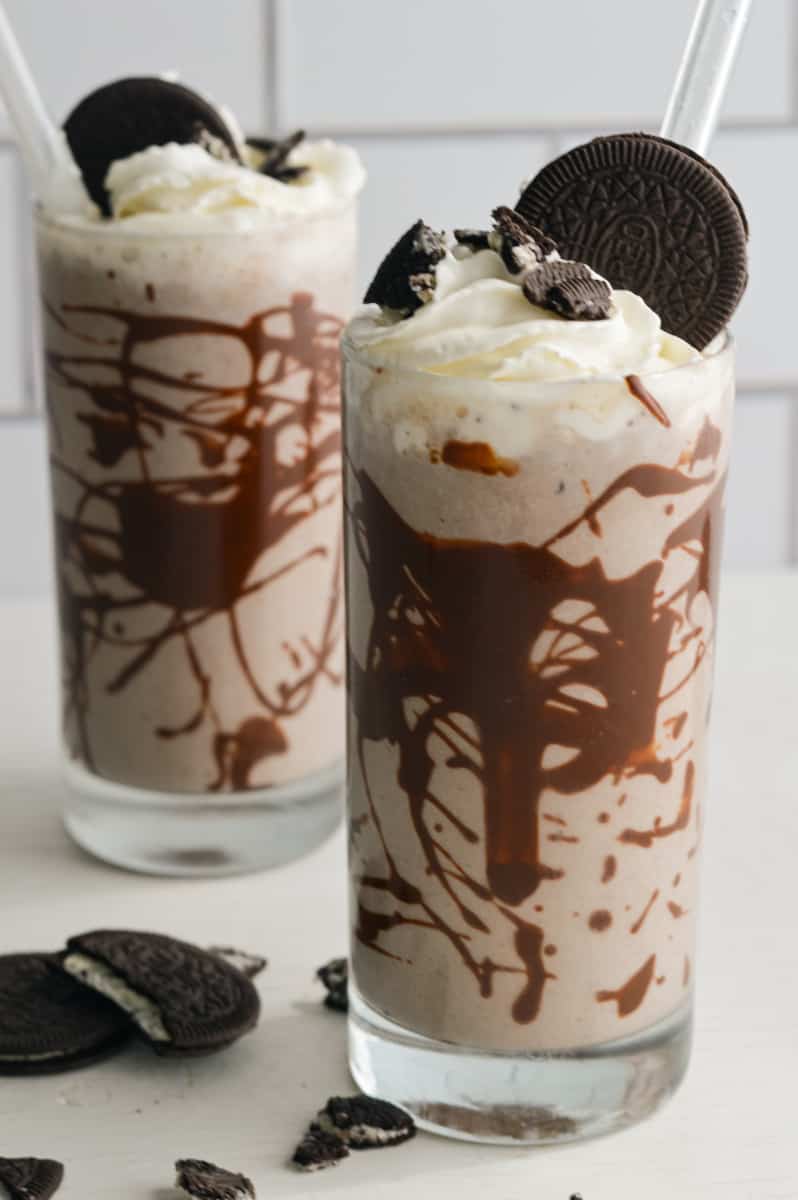 Two Oreo protein shakes in tall glasses topped with whipped cream and cookie crumbs.