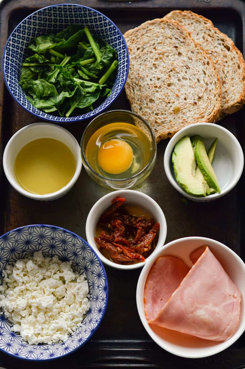 Ingredients for breakfast sandwich including eggs, bacon, veggies, feta and hummus.