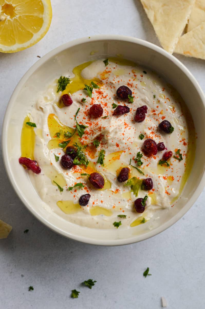 Bowl of tahini yogurt sauce topped with olive oil, pomegranate seeds, parsley and chili powder.