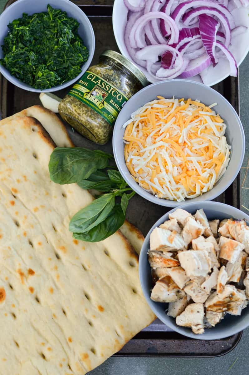 Ingredients including cheese, chicken, red onion, basil, flatbread, and pesto.