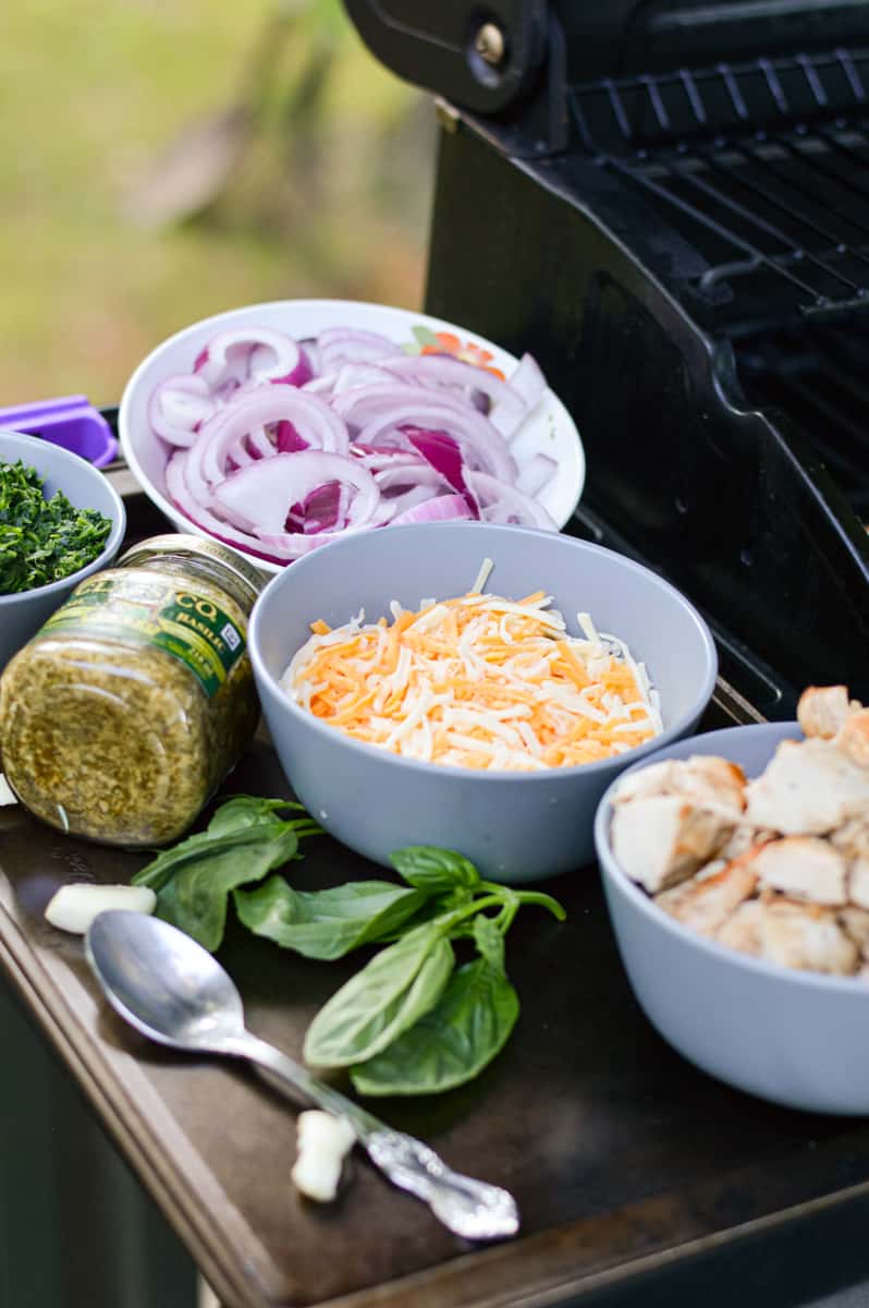 Toppings by the barbecue, including cheese, chicken, basil, red onion and pesto.