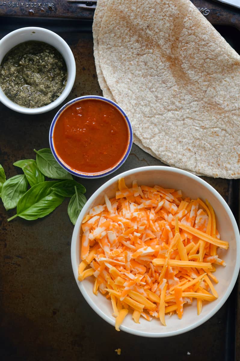 Ingredients including whole wheat tortilla, pesto, tomato sauce, cheese, and basil.