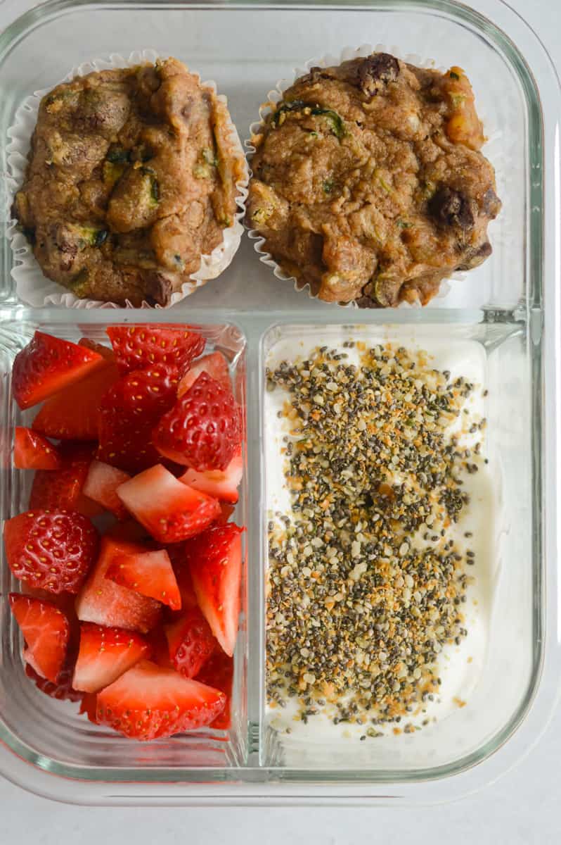 Packed breakfast box with zucchini muffins, yogurt with seeds and strawberries.