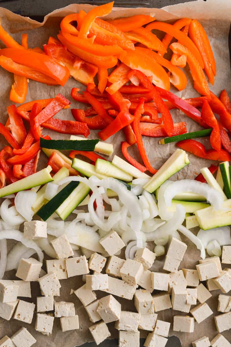 Adding bell peppers, onion, zucchini and cubed tofu to a sheet pan.