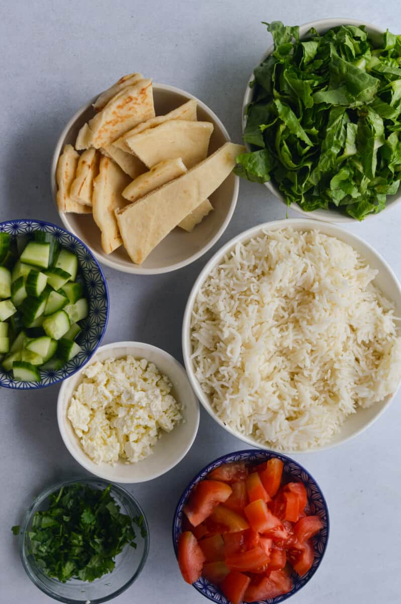 Toppings for chicken shawarma bowls including rice, pita, cucumber, tomatoes, feta and cilantro.