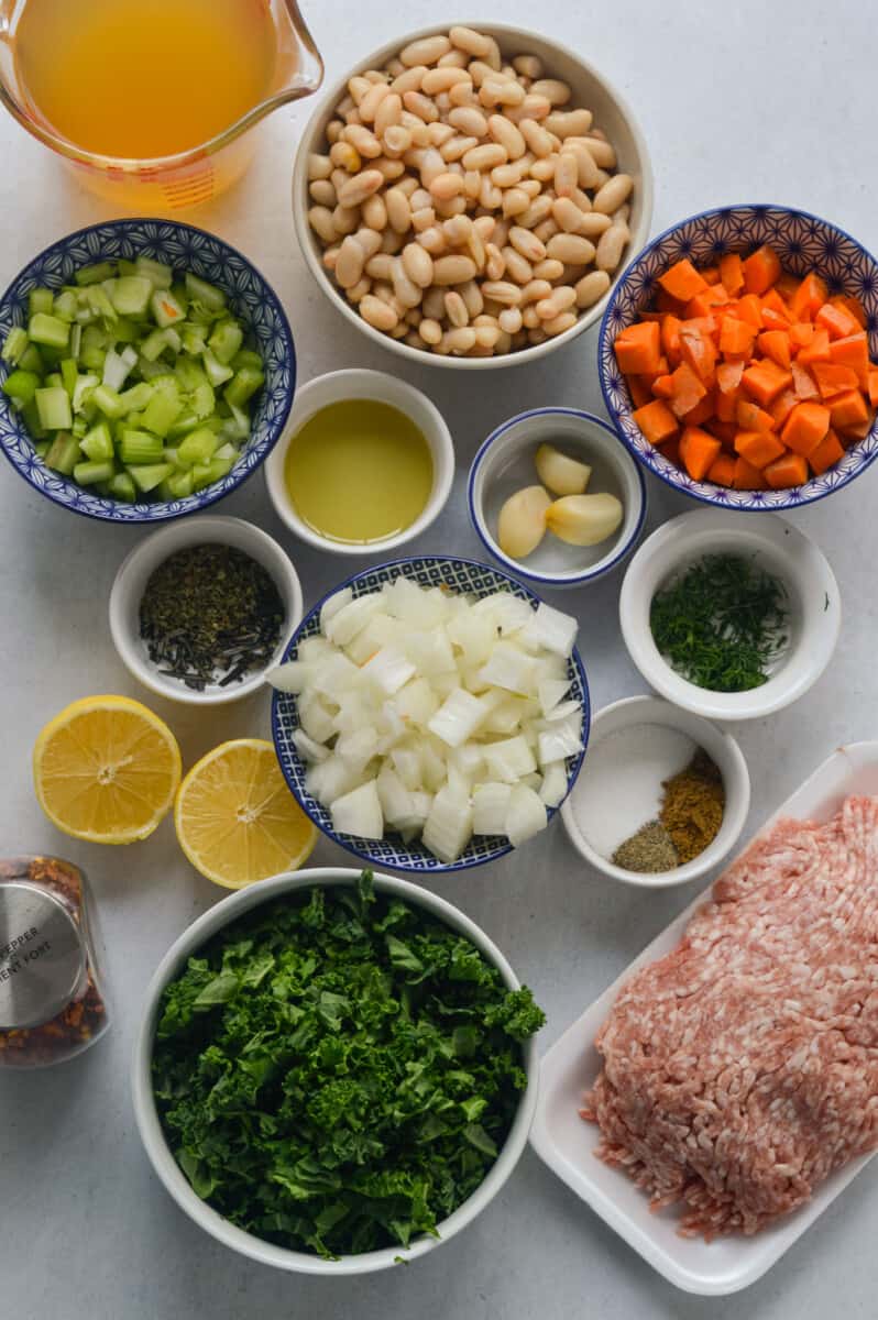Portioned ingredients including ground turkey, diced onions, diced carrots, diced celery, olive oil, spices, kale, parmesan, white beans, garlic and lemon.