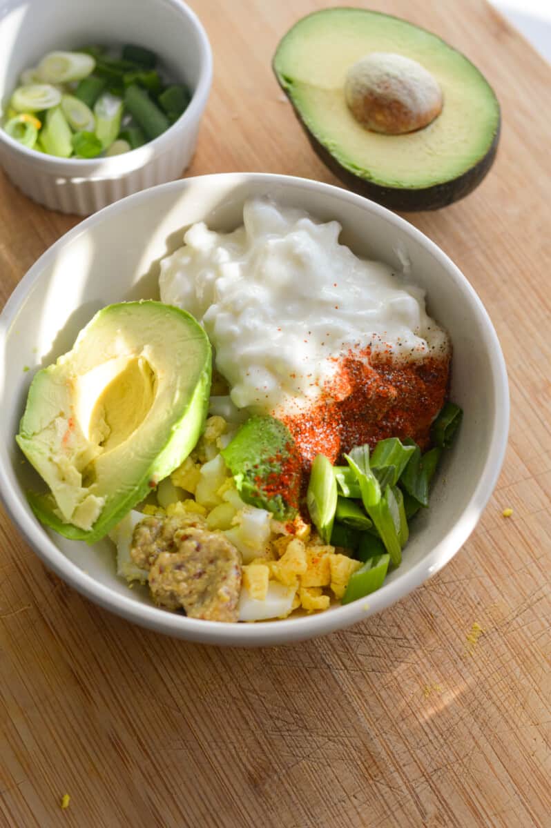 Adding avocado, cottage cheese, spices, dijon mustard and a hard boiled egg into a bowl.