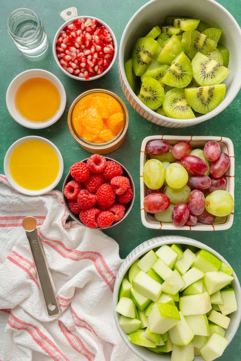 Ingredients for fruit salad including green apples, grapes, cinnamon, raspberries, pomegranate, honey, orange juice and cinnamon