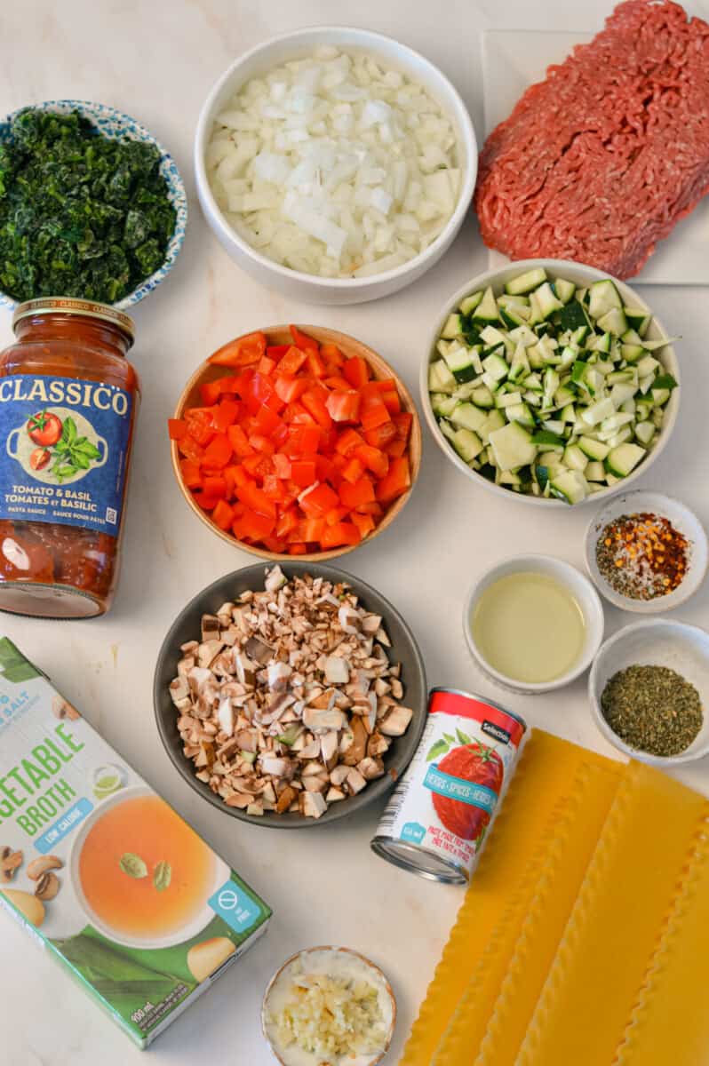 Ingredients for lasagna soup including olive oil, onion, garlic cloves, ground beef, vegetable broth, tomato sauce, tomato paste, spices, cremini mushrooms, zucchini, red bell pepper, lasagna noodles, frozen spinach and parmesan cheese
