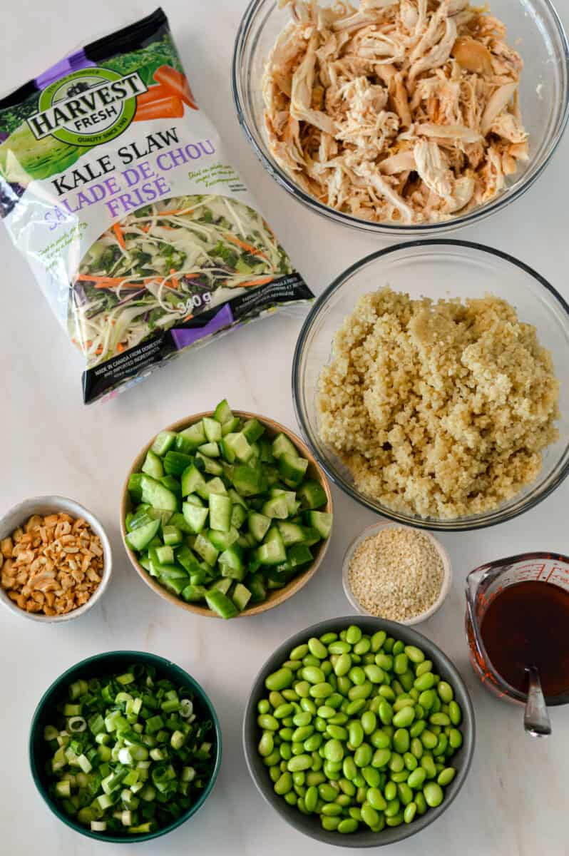 Ingredients for chicken salad including rotisserie chicken, edamame, quinoa, green onions, cashews, sesame seeds, cucumbers, kale slaw, and spicy soy dressing