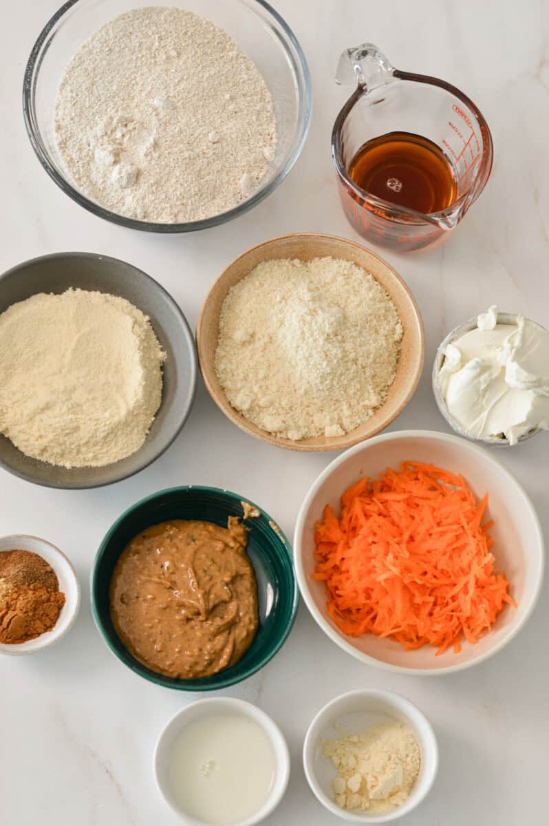 Ingredients for no-bake protein bars including oat flour, almond flour, vanilla protein powder, spices, nut butter, maple syrup, shredded carrots, cream cheese and vanilla protein powder