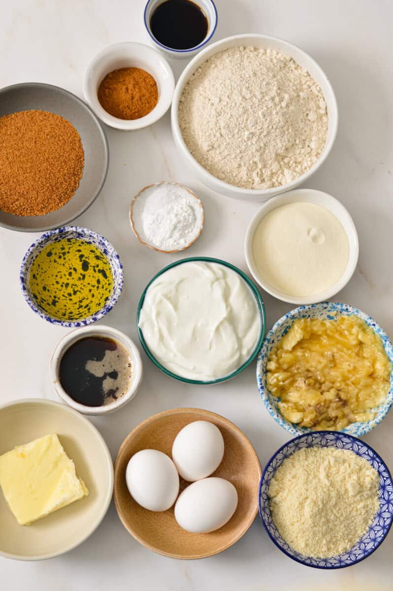 Ingredients for coffee cake including oat flour, protein powder, leavening agents, plain Greek yogurt, mashed bananas, maple syrup, cinnamon, salt, olive oil, eggs, vanilla extract, ingredients for oat crumble topping and white chocolate