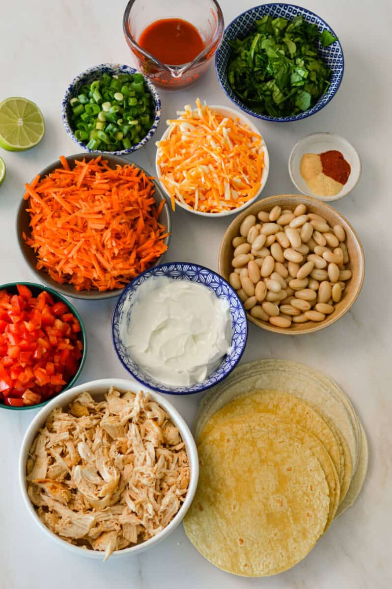 Ingredients for the chicken taquitos including tortillas, cheese, chicken, veggies, buffalo sauce, spices, white beans, Greek yogurt, lime, and green onions