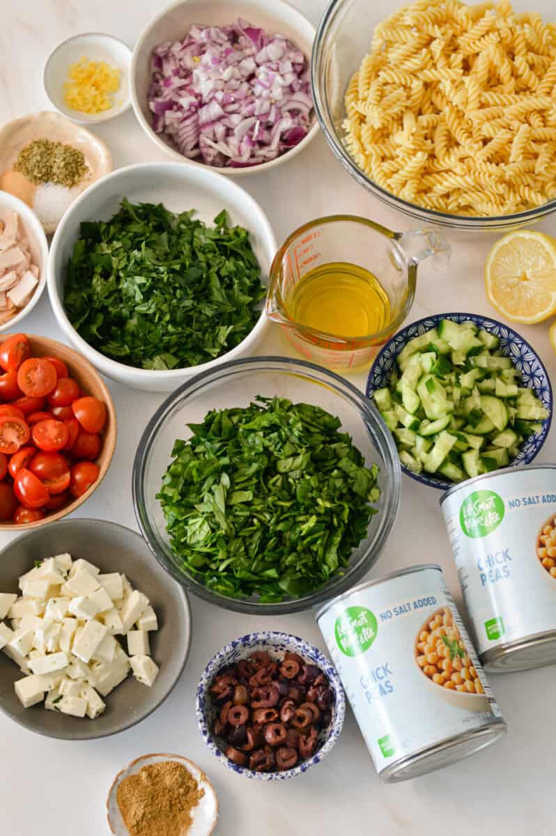 Ingredients for beans pasta salad including chickpeas, feta, cucumber, parsley, tomatoes, deli turkey, red onion, black olives, lemon, spinach and spices.