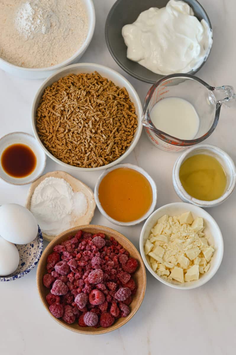 Ingredients for bran muffins including frozen raspberries, Greek yogurt, all-bran cereal, white chocolate, honey, whole wheat pastry flour, eggs, olive oil, leavening agents, and vanilla.