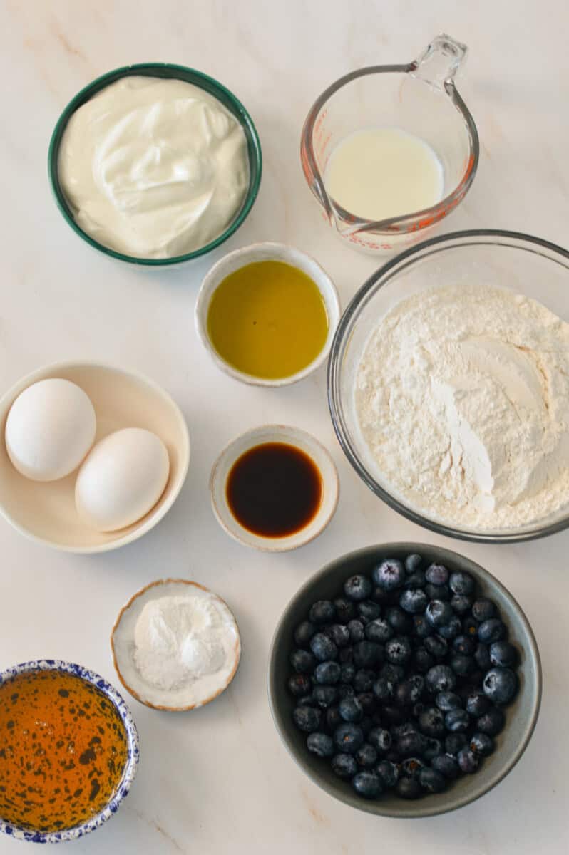 Ingredients for yogurt muffins including Greek yogurt, milk, vanilla extract, olive oil, honey, flour, leavening agents, salt, and blueberries.