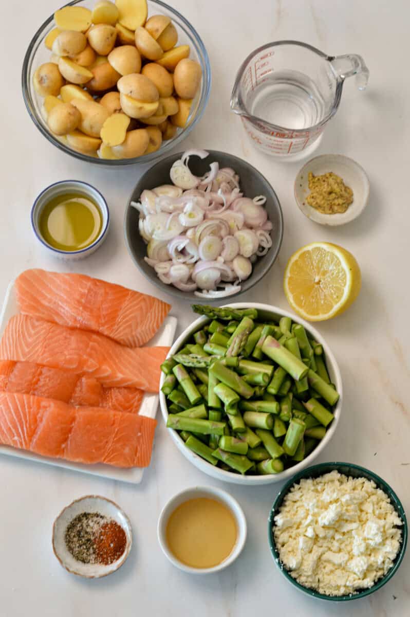 Ingredients for sheet pan dinner including salmon, lemon, dill, baby potatoes, asparagus, shallot, butter, garlic, dijon mustard, olive oil, spices, water and feta.