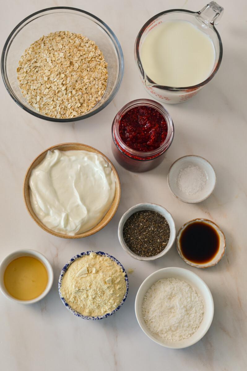 Ingredients for raspberry overnight oats including oats, Greek yogurt, raspberry chia jam, vanilla protein powder, chia seeds, vanilla, honey, coconut milk, and unsweetened shredded coconut.