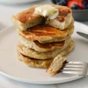 Stack of banana cottage cheese pancakes on a plate with butter. A fork has taken a bite out of it.