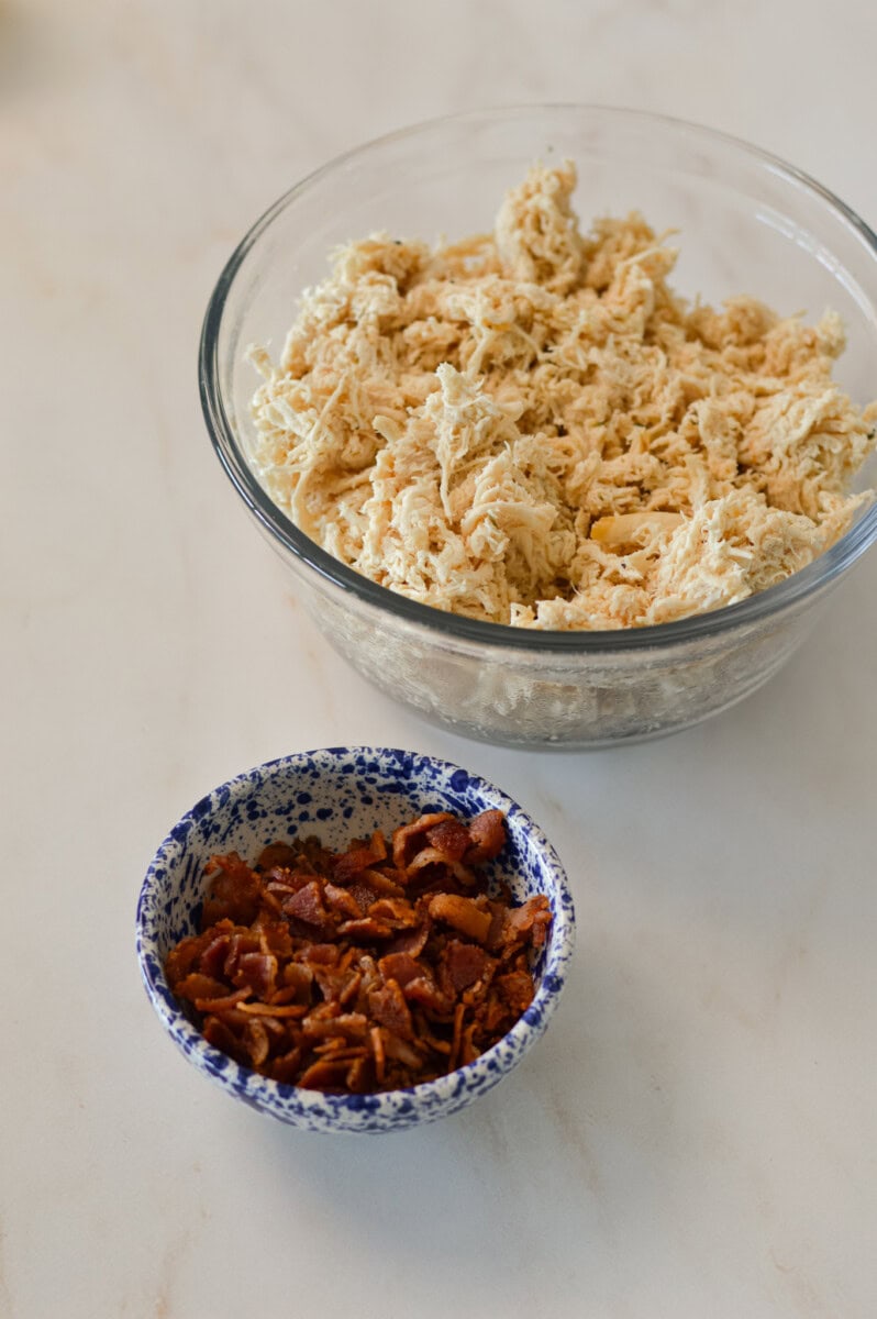 Shredded cooked chicken in a bowl. Cooked bacon in small pieces in a bowl.