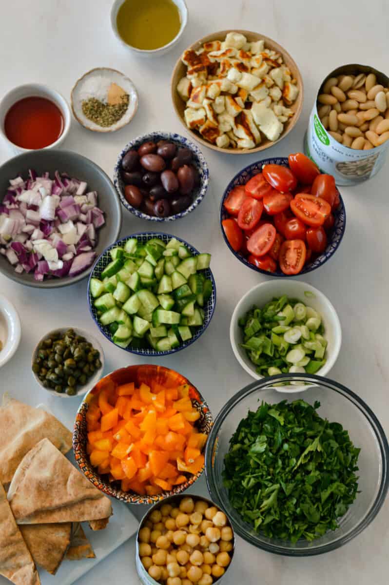 Ingredients for dense bean salad including halloumi, chickpeas, white beans, cherry tomatoes, cucumbers, red onion, kalamata olives, capers, orange bell pepper, green onions, pitas, fresh herbs, dijon mustard, red wine vinegar, and spices.