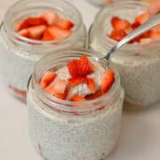 Yogurt chia pudding in jars topped with strawberries. A spoon is lifting up a bite out of one of the jars.