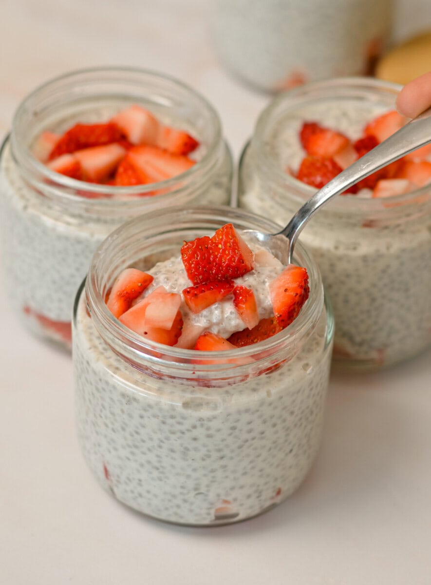 Yogurt chia pudding in jars topped with strawberries. A spoon is lifting up a bite out of one of the jars.