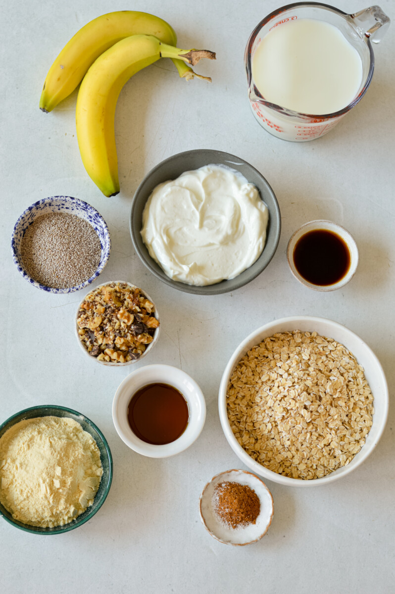 Ingredients for overnight oats including bananas, Greek yogurt, oats, vanilla protein powder, milk, vanilla extract, maple syrup, walnuts, chocolate chips, and cinnamon.