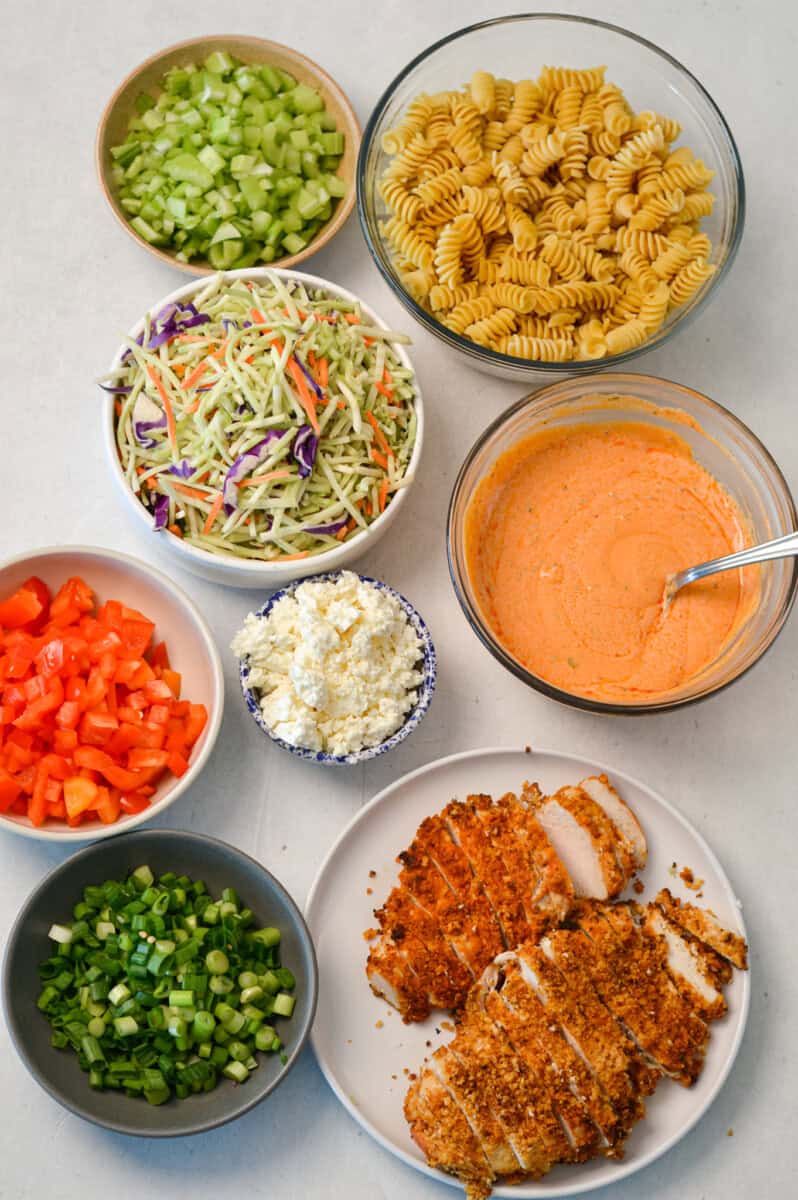 Ingredients for the protein pasta salad including buffalo sauce, crispy chicken, feta, green onions, protein pasta, kale slaw, celery, red bell pepper, and Greek yogurt buffalo sauce.
