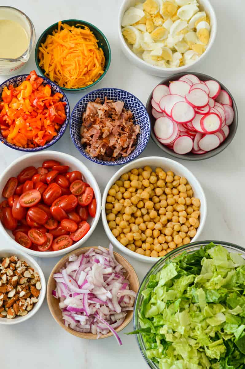 Ingredients for layered salad including romaine lettuce, tomatoes, chickpeas, cheddar cheese, radishes, hard-boiled eggs, bacon, red onion, bell pepper and creamy honey mustard dressing.