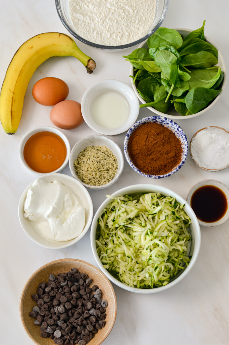 Ingredients for veggie muffins inclufing banana, spinach, zucchini, honey, plain yogurt, egg, milk, hemp hearts, all purpose flour, cocoa powder, baking powder, baking soda, salt, and chocolate chips.