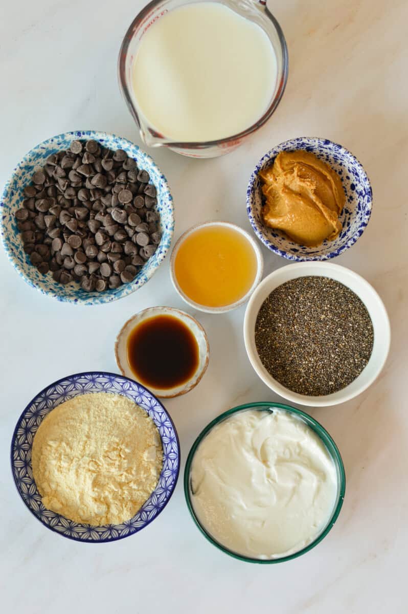 Ingredients for chocolate chia pudding including chocolate chips, peanut butter, Greek yogurt, vanilla protein powder, chia seeds, milk, honey, and vanilla extract.