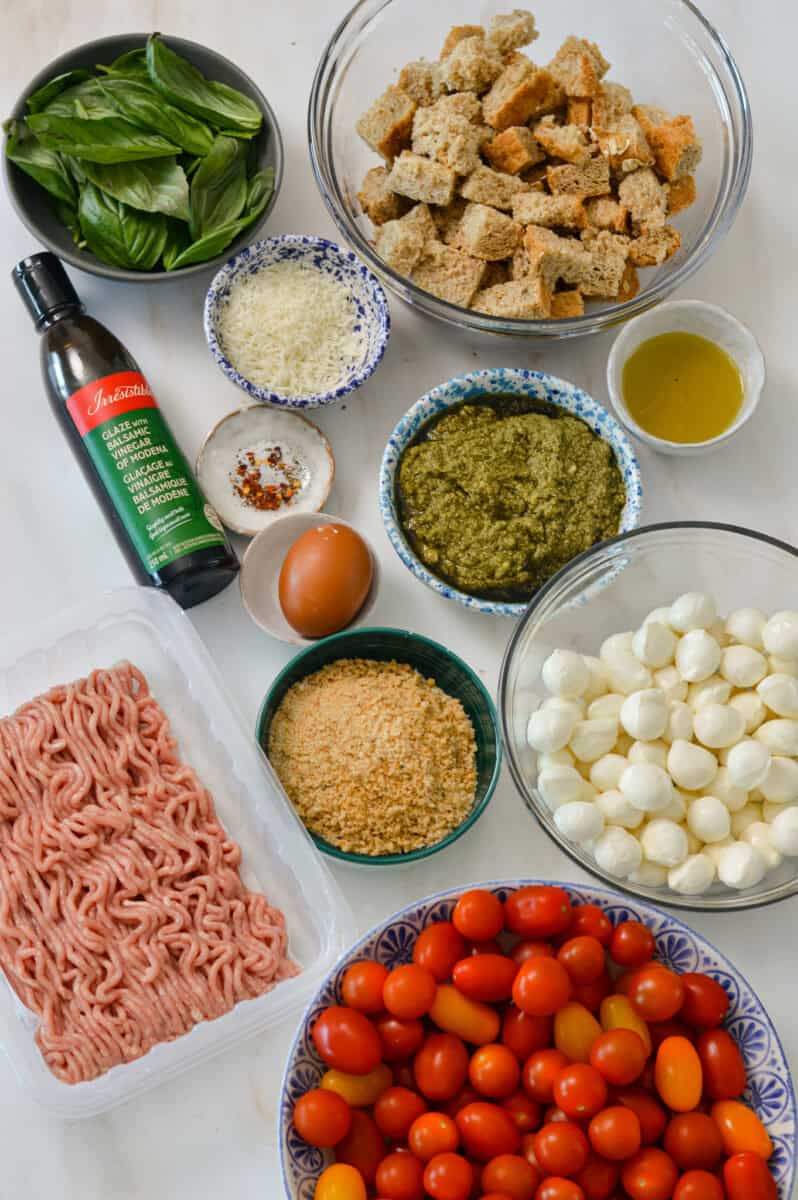 Ingredients for pesto chicken meatballs including ground chicken, breadcrumbs, bocconcini, pesto, cherry tomatoes, olive oil, basil, balsamic glaze, and sourdough bread.