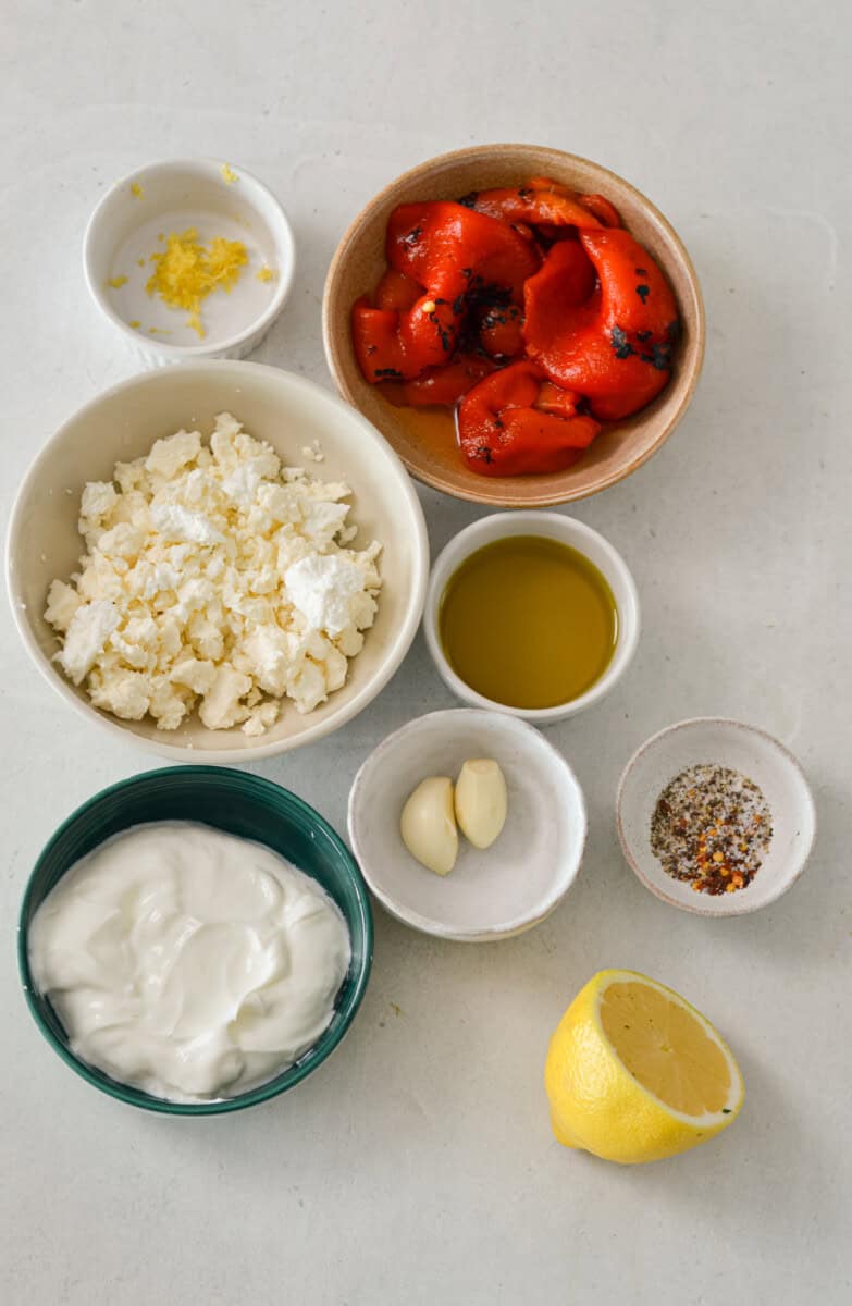 Ingredients for red pepper feta dip including roasted red peppers, feta, garlic, lemon, Greek yogurt, lemon zest, spices, and olive oil.