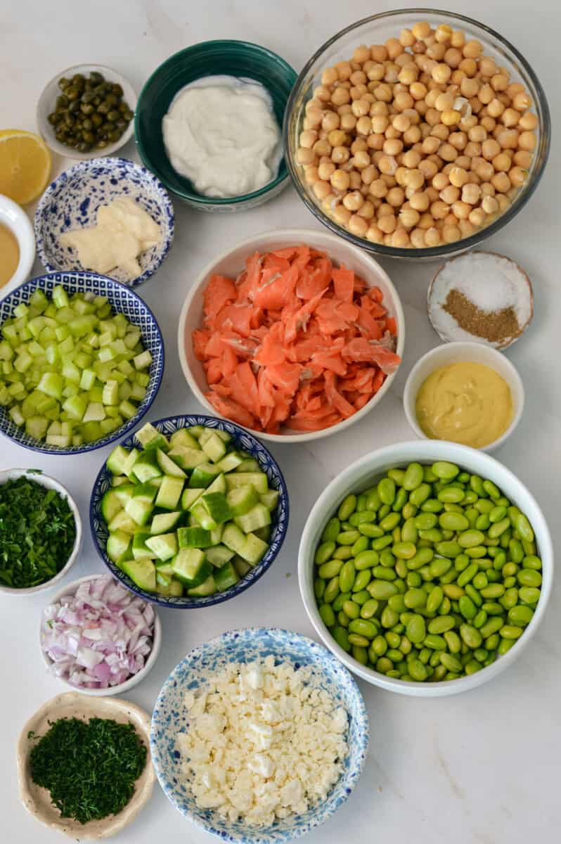 Ingredients for salad including chickpeas, feta, cucumber, edamame, fresh dill, fresh parsley, smoked salmon, cumin, Dijon mustard, lemon juice, shallot, celery, Greek yogurt and mayonnaise.
