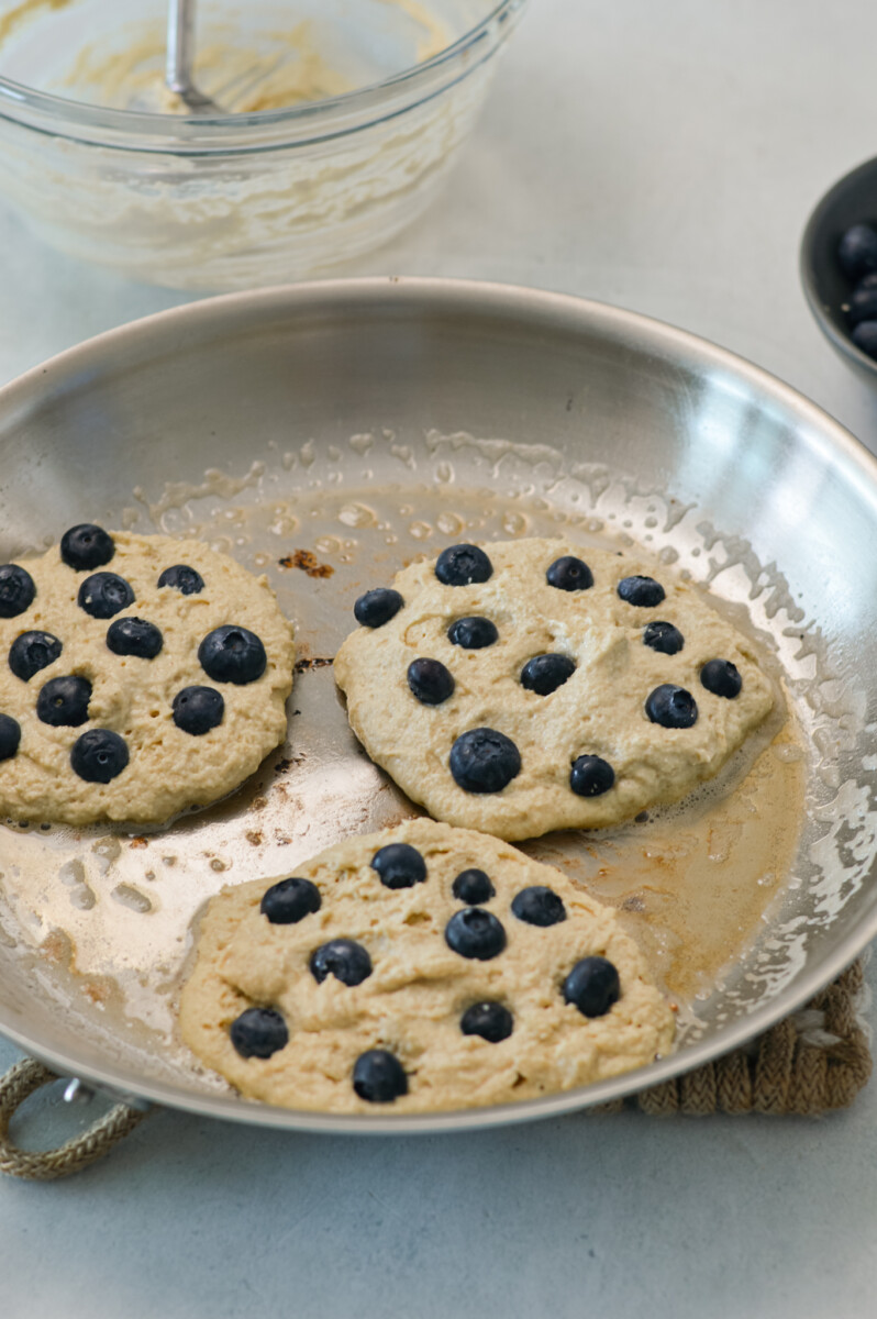 Pancakes cooking on a pan, topped with blueberries.