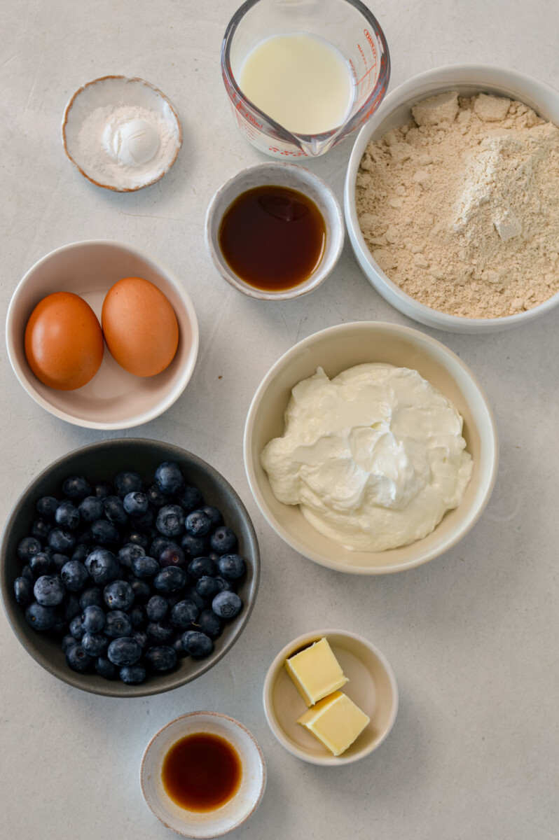 Ingredients for blueberry pancakes including blueberries, Greek yogurt, oat flour, eggs, butter, vanilla extract, maple syrup, baking powder, and salt.