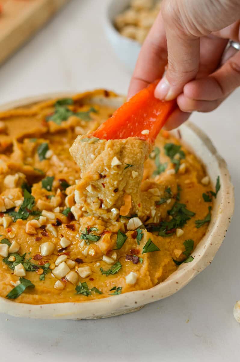 Red pepper scooping up peanut butter hummus out of a bowl.