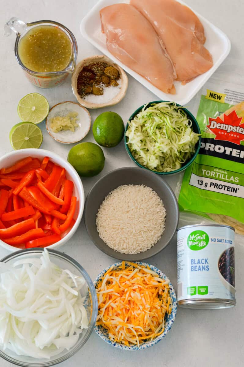 Ingredients for Green Chili Chicken Burritos including chicken, bell peppers, salsa verde, zucchini, rice, onion, protein wraps, black beans, cheese, limes, and zucchini.