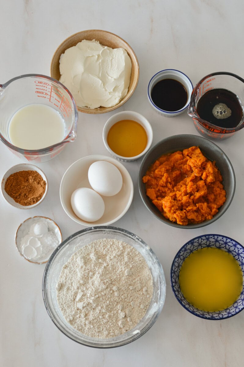 Ingredients for pumpkin muffins including pumpkin puree, eggs, milk, all-purpose flour, oat flour, pumpkin pie spice, honey, maple syrup, cream cheese, vanilla, butter, baking powder and baking soda, and pumpkin seeds.