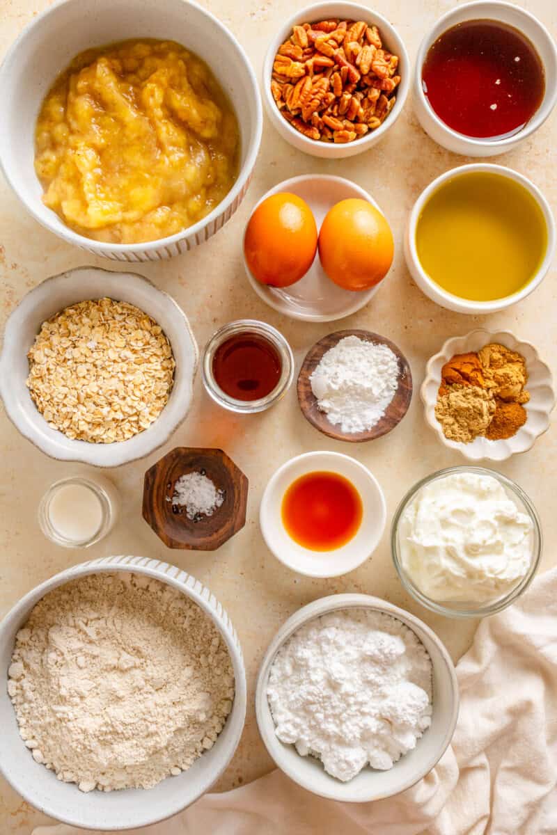 Ingredients for chai banana bread including oat flour, rolled oats, baking powder, baking soda, ground cinnamon, ground cardamom, ground ginger, ground cloves, salt, chopped pecans, bananas, eggs, olive oil, plain Greek yogurt, maple syrup, chai, vanilla extract, icing sugar, and milk.