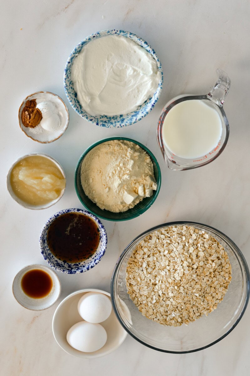 Ingredients for baked oats including oats, eggs, milk, maple syrup, vanilla extract, vanilla protein powder, cinnamon, baking powder, salt, butter, sugar, plain Greek yogurt, and maple syrup.